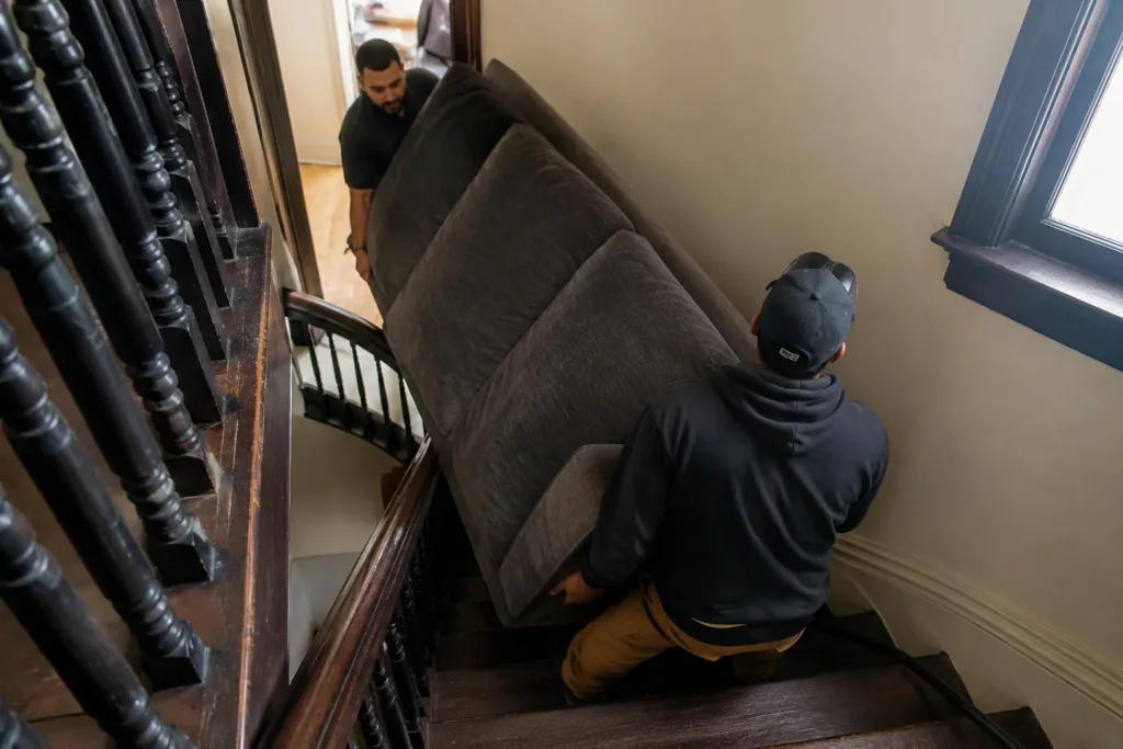 DIY Junk Removal vs Hiring a Pro in New Bedford — How to Decide 2 Professional movers carrying a large sofa down a narrow triple-decker staircase during a furniture removal job in New Bedford, Massachusetts.